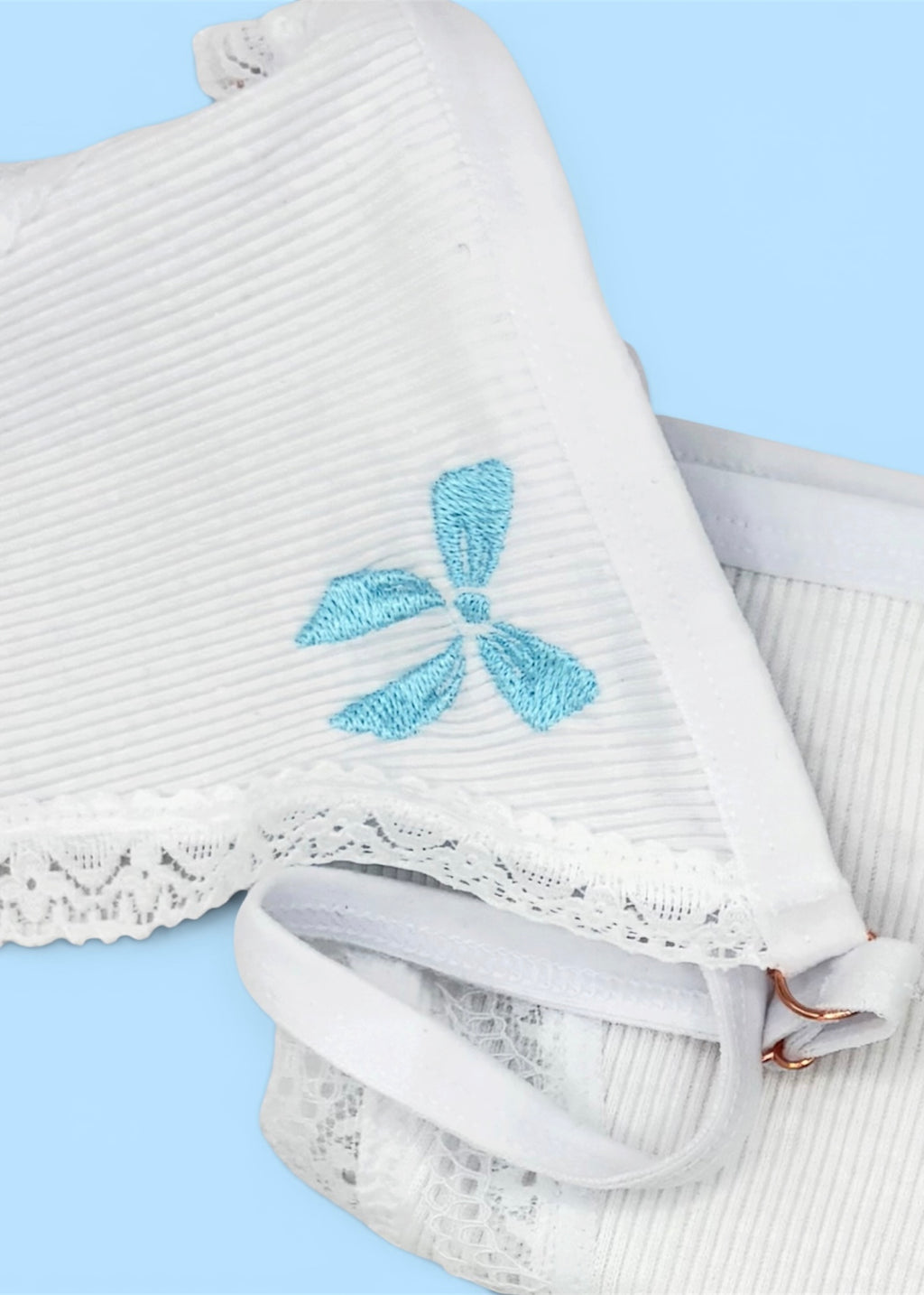 A close up image of Berry Sorbet’s Sugar Bow thong over a light blue background that accentuates the light blue bow embroidery on the front of the white ribbed cotton thong.
