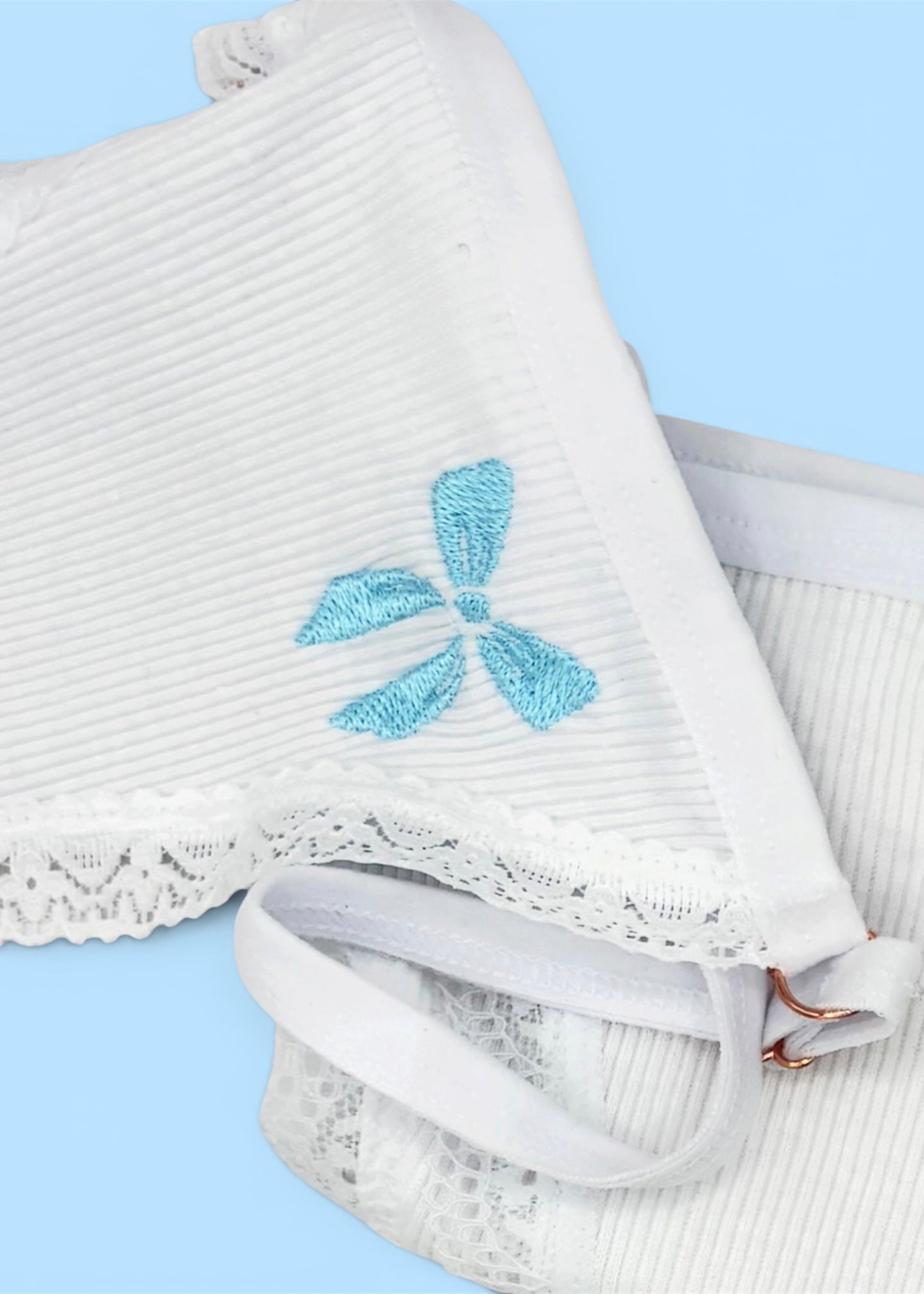 A close up image of Berry Sorbet’s Sugar Bow thong over a light blue background that accentuates the light blue bow embroidery on the front of the white ribbed cotton thong.